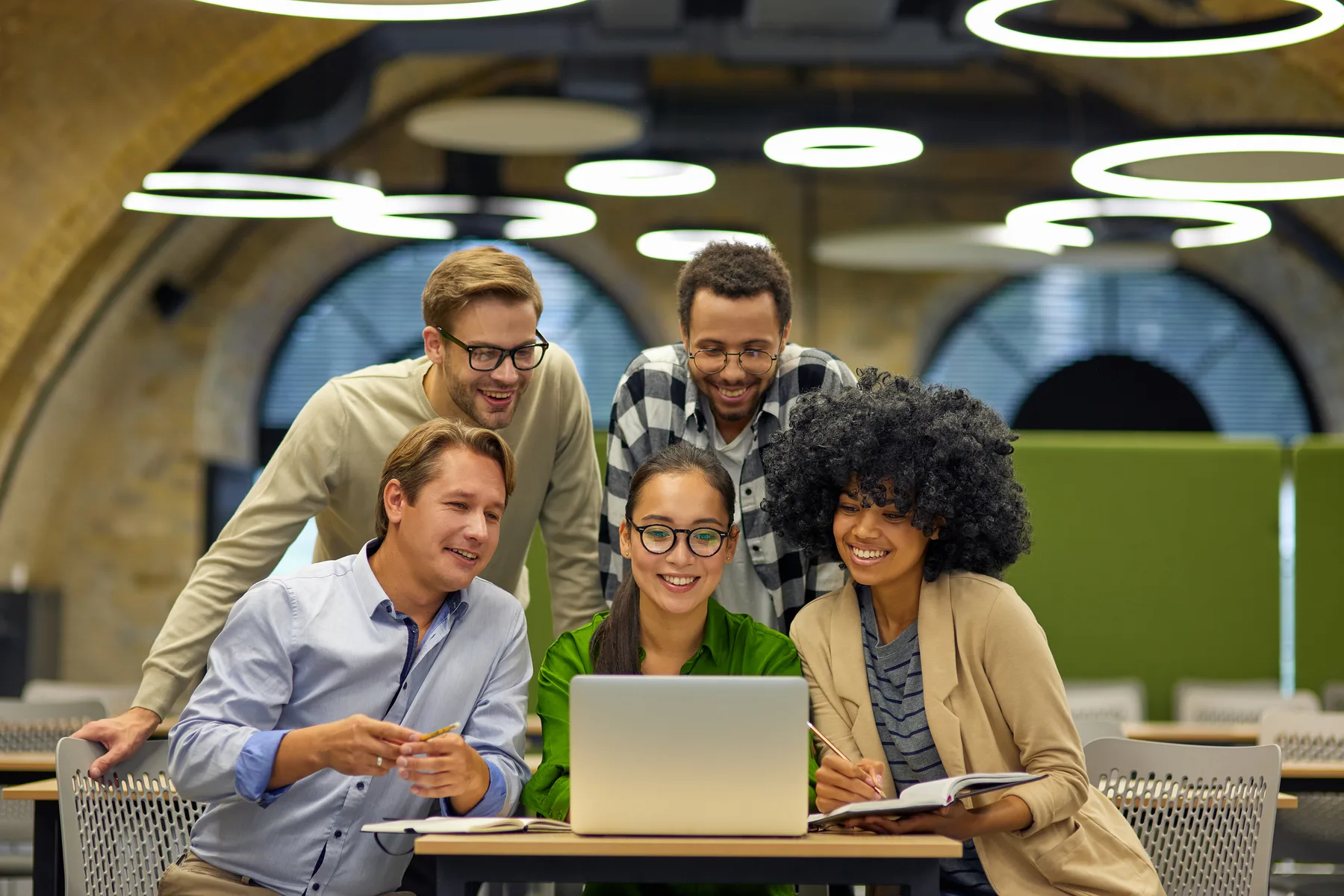A five-person small business team gathered around a laptop in a modern coworking space, reviewing work together and smiling