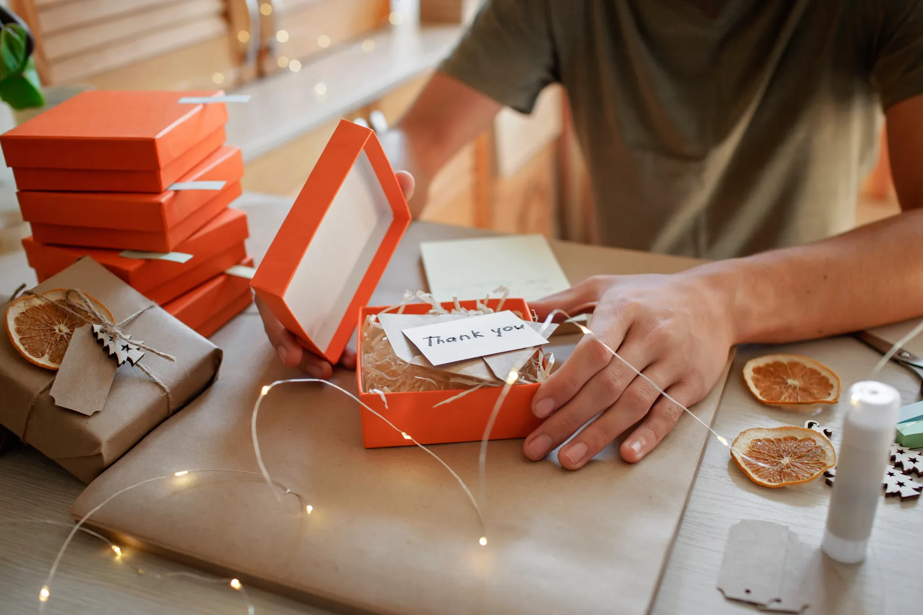 A small business owner packing an orange gift box with a handwritten thank-you note on a table decorated with string lights