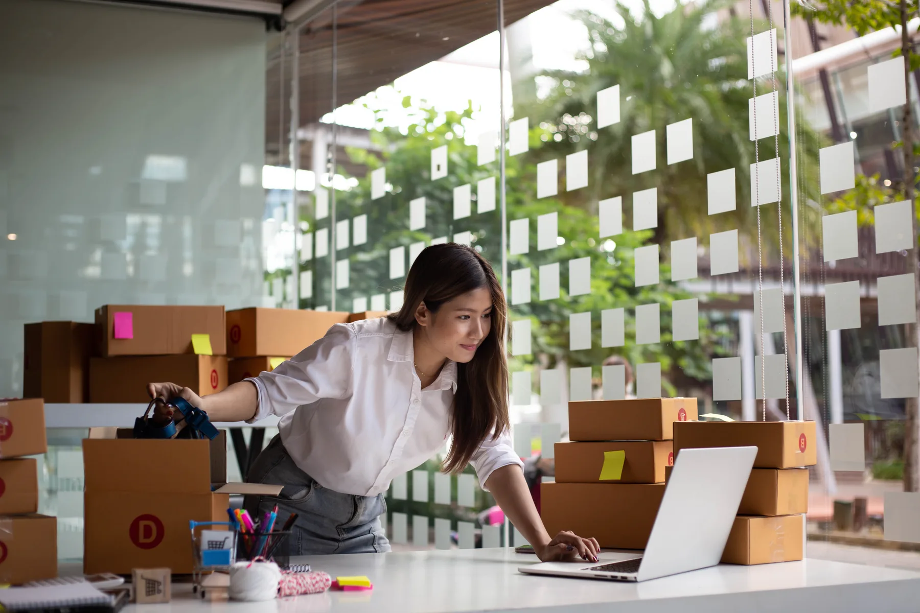 A small business owner reviewing orders on a laptop surrounded by labeled shipping boxes