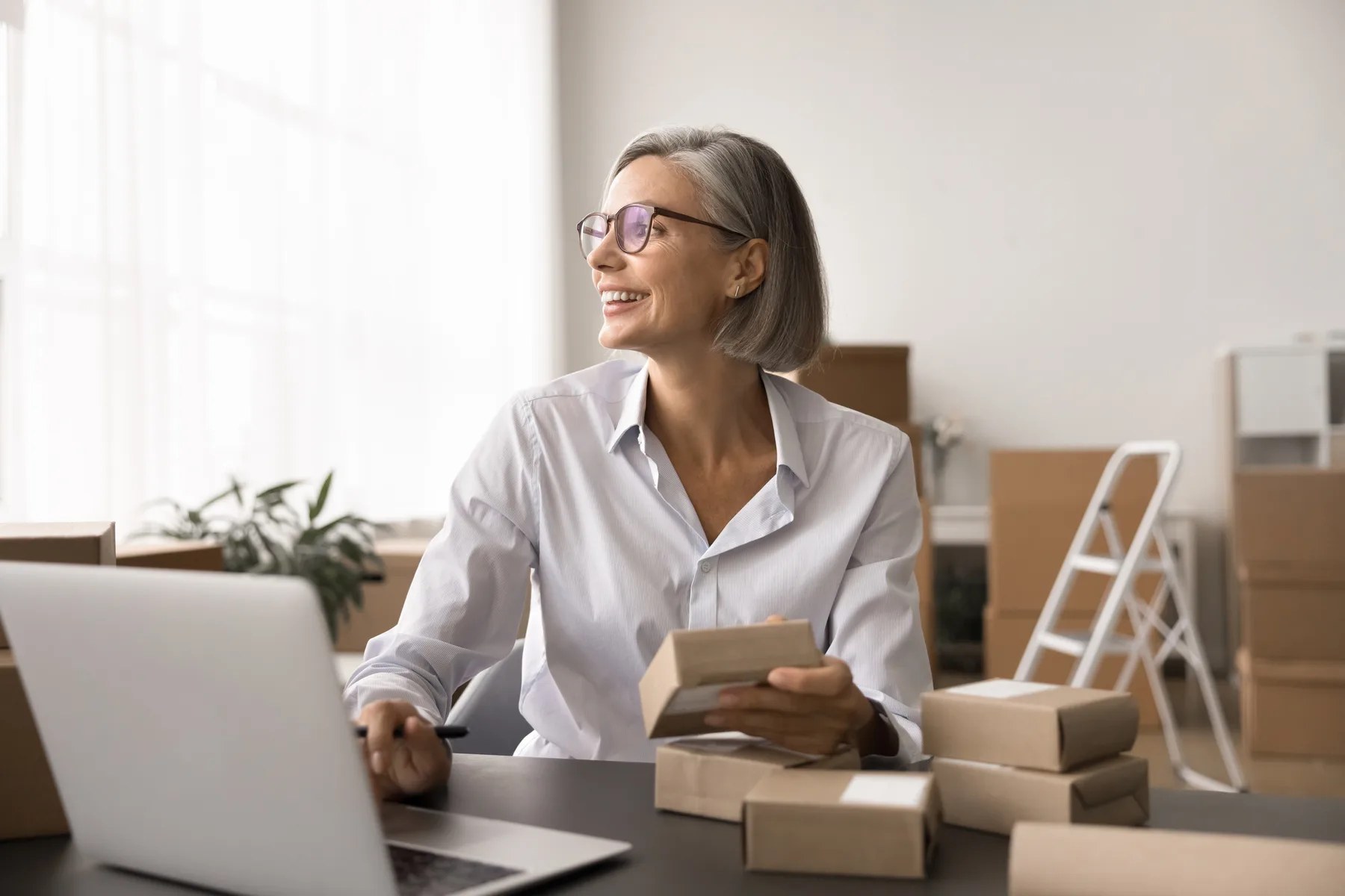 A smiling retail business owner working on her laptop at a desk surrounded by packed shipping boxes