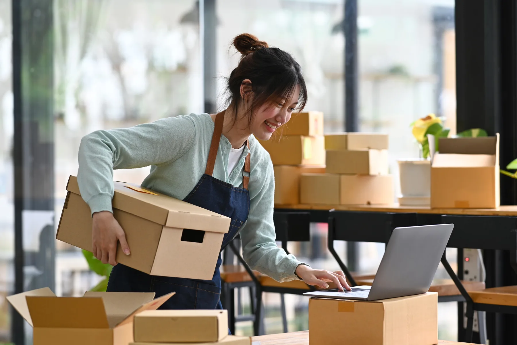An online store owner checking an order on her laptop while holding a packed shipping box